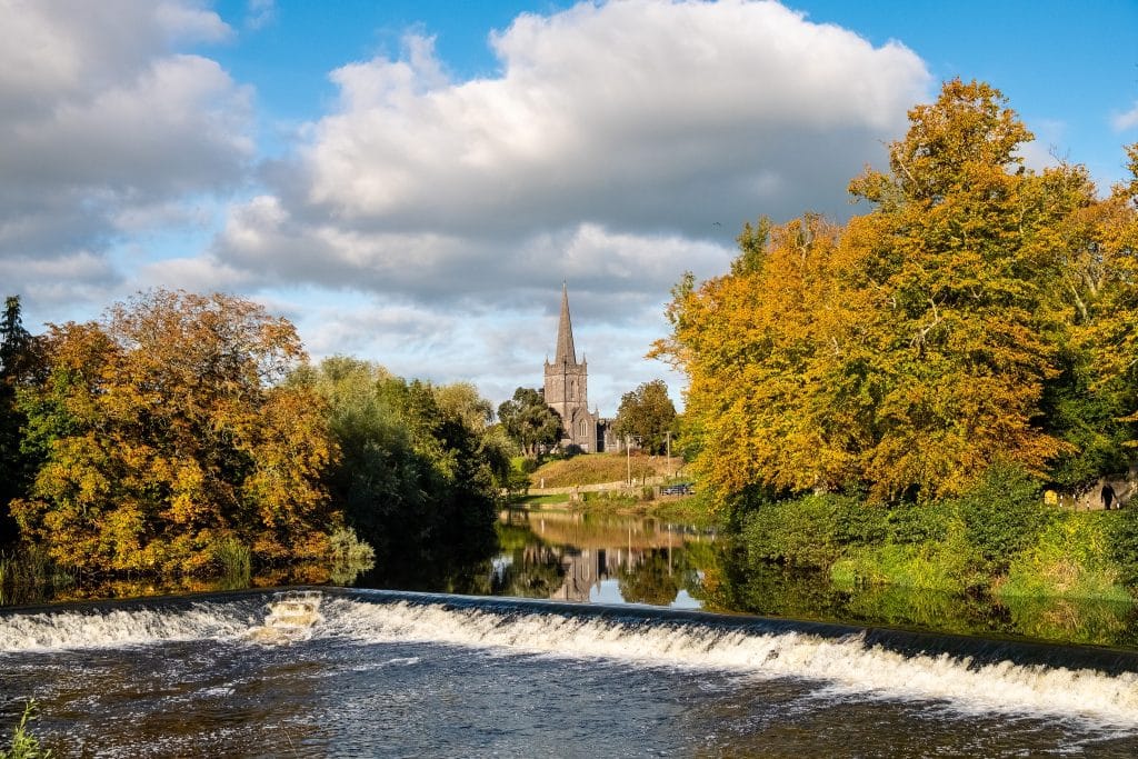A serene river scene with a small waterfall in the foreground, flanked by trees with autumn foliage. In the background stands a tall church steeple under a blue sky with scattered clouds, reflecting on the calm water.