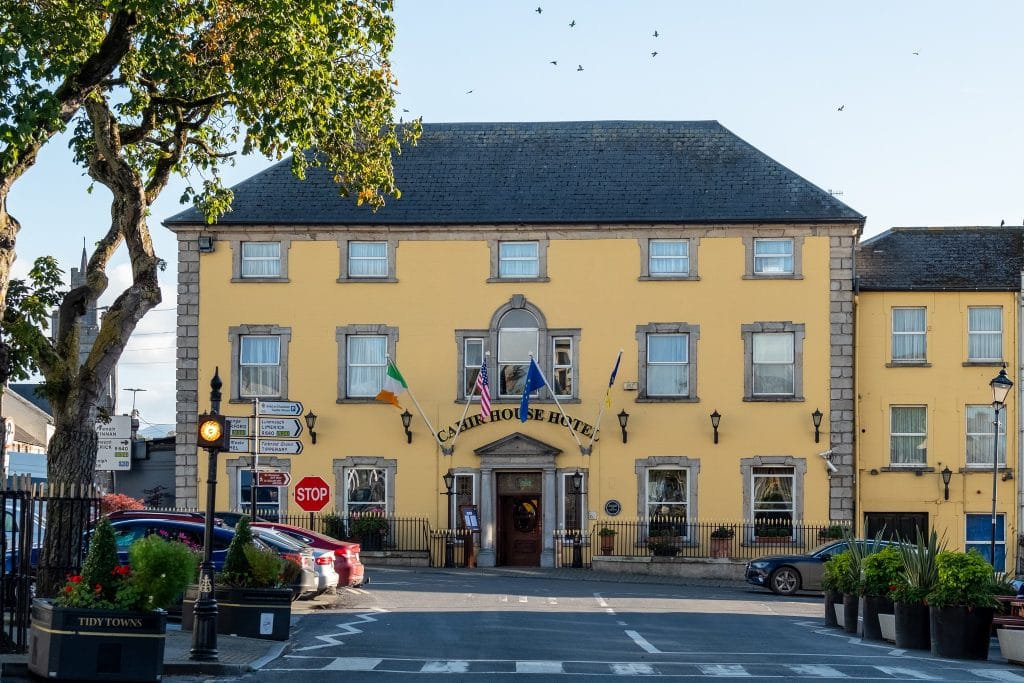 A yellow two-story building with a slate roof, identified as the Castle House Hotel, displaying the Irish, American, and European Union flags above the entrance. In front, a street with a stop sign, directional road signs, parked cars, and decorated planters. A tree with green leaves is on the left side, and birds are flying in the clear sky.