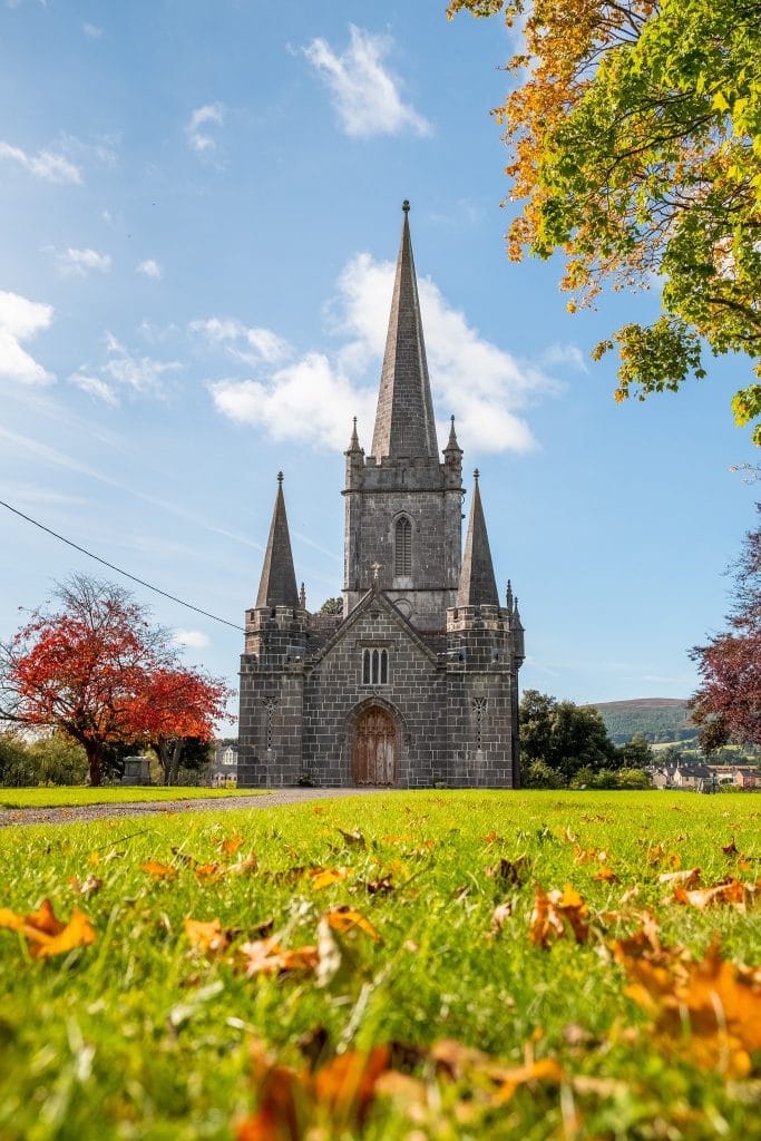 Stone church with three tall spires on a grassy lawn scattered with autumn leaves, under a blue sky with some clouds.