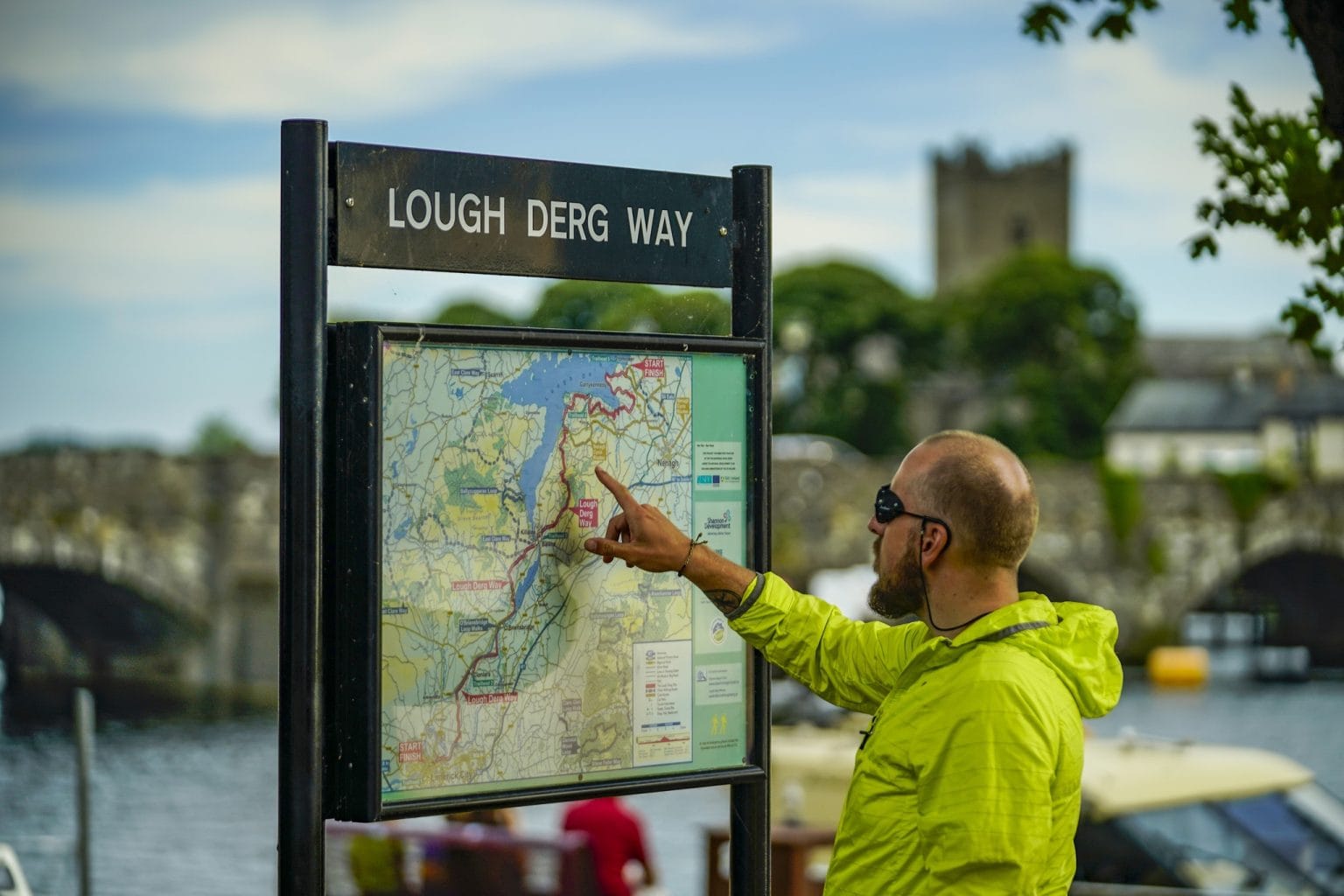 A man wearing a bright yellow jacket and sunglasses is pointing at a map on a black signpost labeled "Lough Derg Way" near a body of water with a stone bridge and buildings in the background.