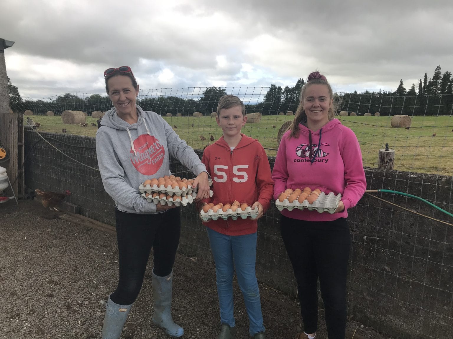 Three people standing outdoors in front of a fenced field holding trays of eggs, with hay bales and chickens visible in the background under a cloudy sky.