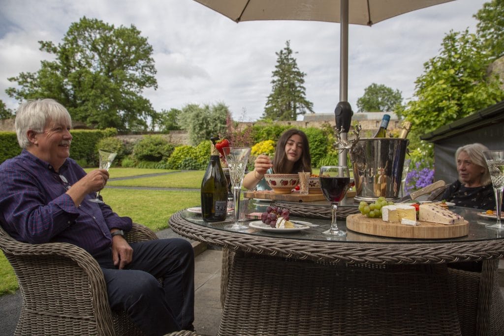 Three people enjoying a relaxed outdoor gathering around a wicker table with wine, cheese, grapes.