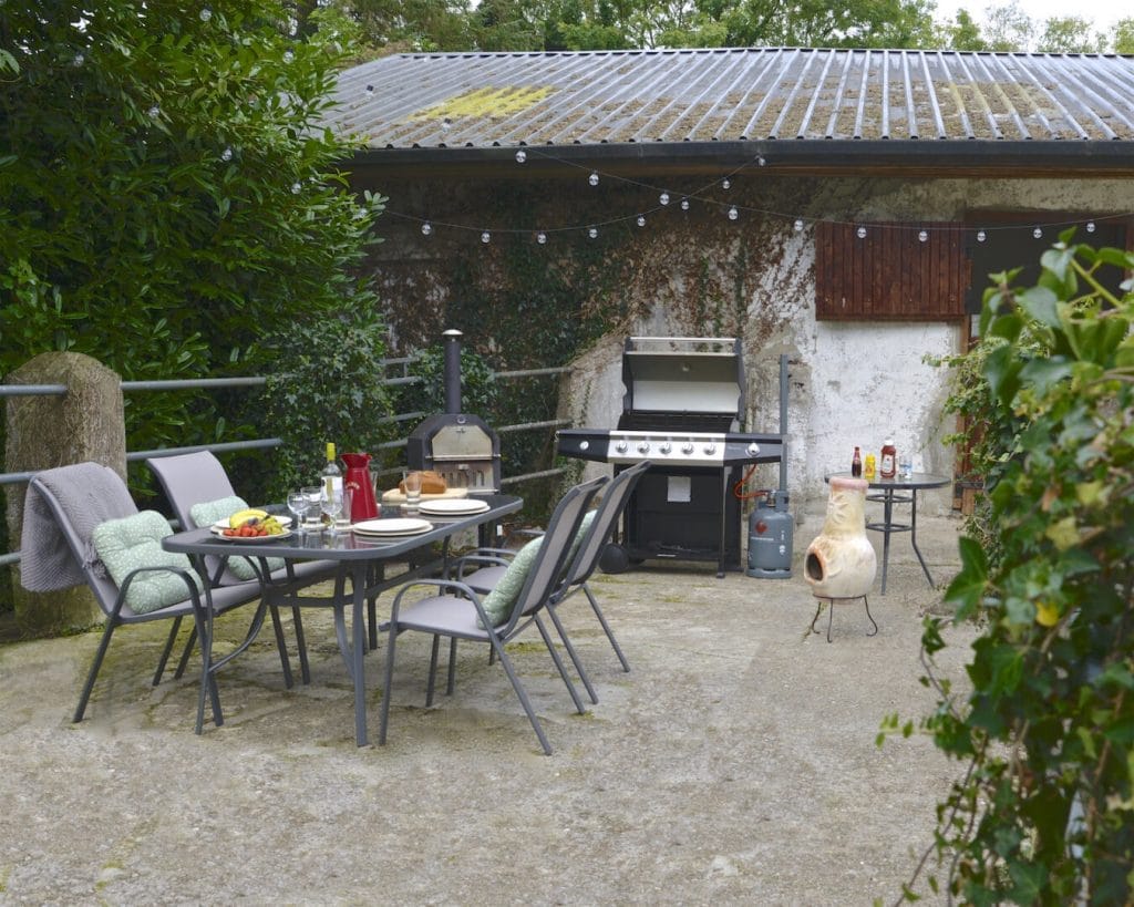 Outdoor patio area with a glass-top dining table set with plates, glasses, and food including fruit and bread. Four cushioned chairs surround the table, two with a blanket draped over the back. In the background, there is a gas grill, a small wood-fired oven, and a clay chiminea. String lights hang overhead, and greenery surrounds the space with trees and bushes. The setting appears cozy and prepared for a meal.