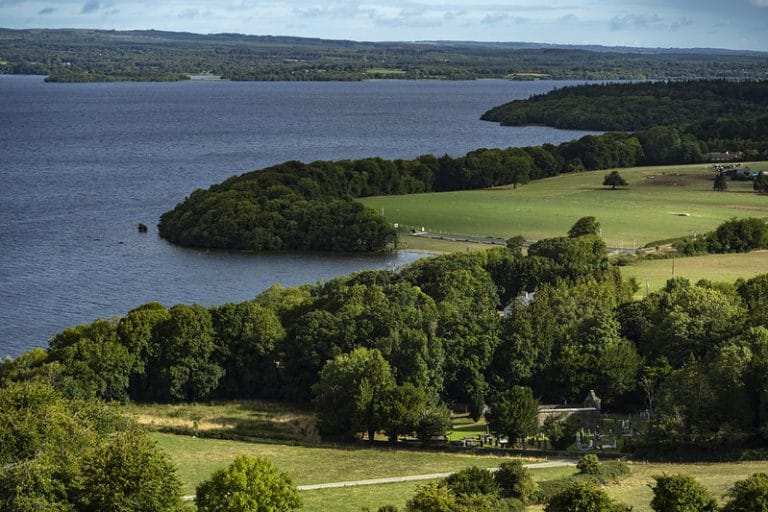 Scenic view of a lake bordered by lush green trees and open grassy fields under a partly cloudy sky.