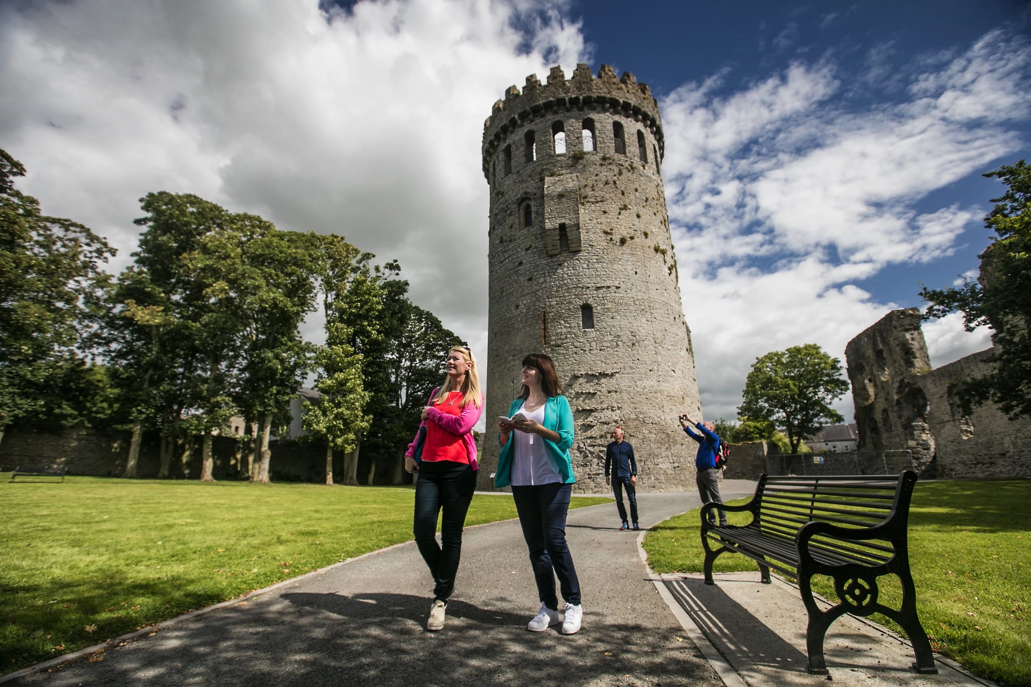 Two women walking and chatting on a path in front of a large, round stone tower with battlements, surrounded by green grass, trees, and ruins, under a partly cloudy sky; a man stands nearby while another takes a photo.