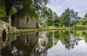 Stone building and lush green trees reflected in a calm pond under a partly cloudy sky.