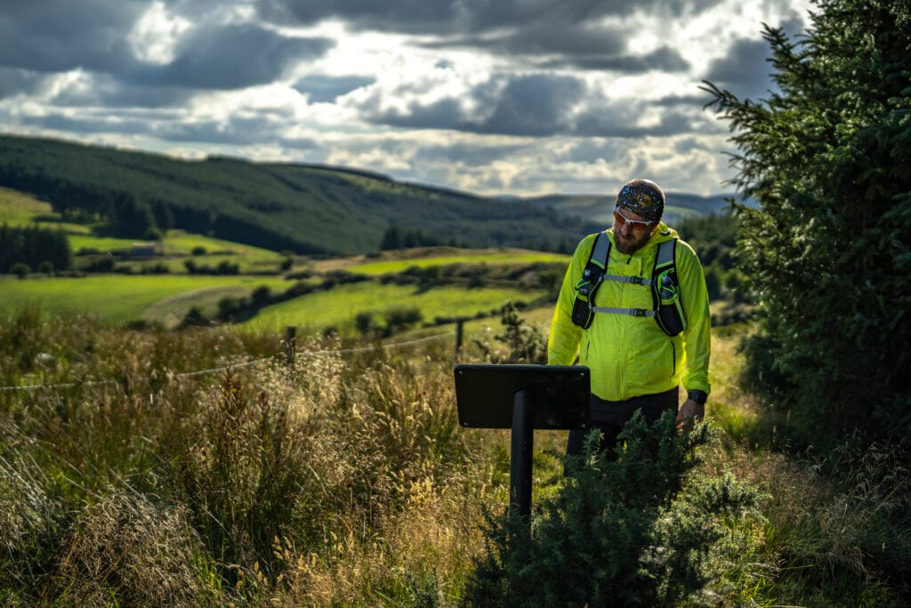 Man in a bright yellow jacket and headband reading an information board along a grassy, rural hiking trail with rolling hills and a cloudy sky in the background.