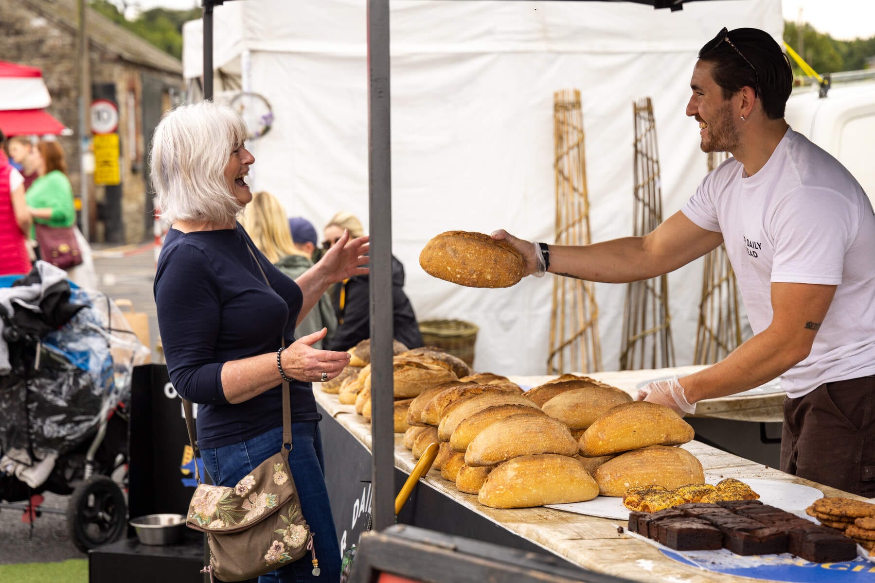 A smiling vendor hands a loaf of bread to a happy older woman at an outdoor market stall filled with various baked goods.