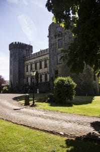 A historic stone castle with two round towers and battlements, set against a clear sky. The building is surrounded by a well-maintained lawn and a curved cobblestone path in the foreground. Tree branches partially frame the right side of the image.