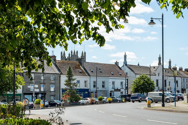 A sunny street scene in a small town with parked cars along a curved road, white and stone buildings with pitched roofs, framed by green tree branches in the foreground and a church tower in the background.