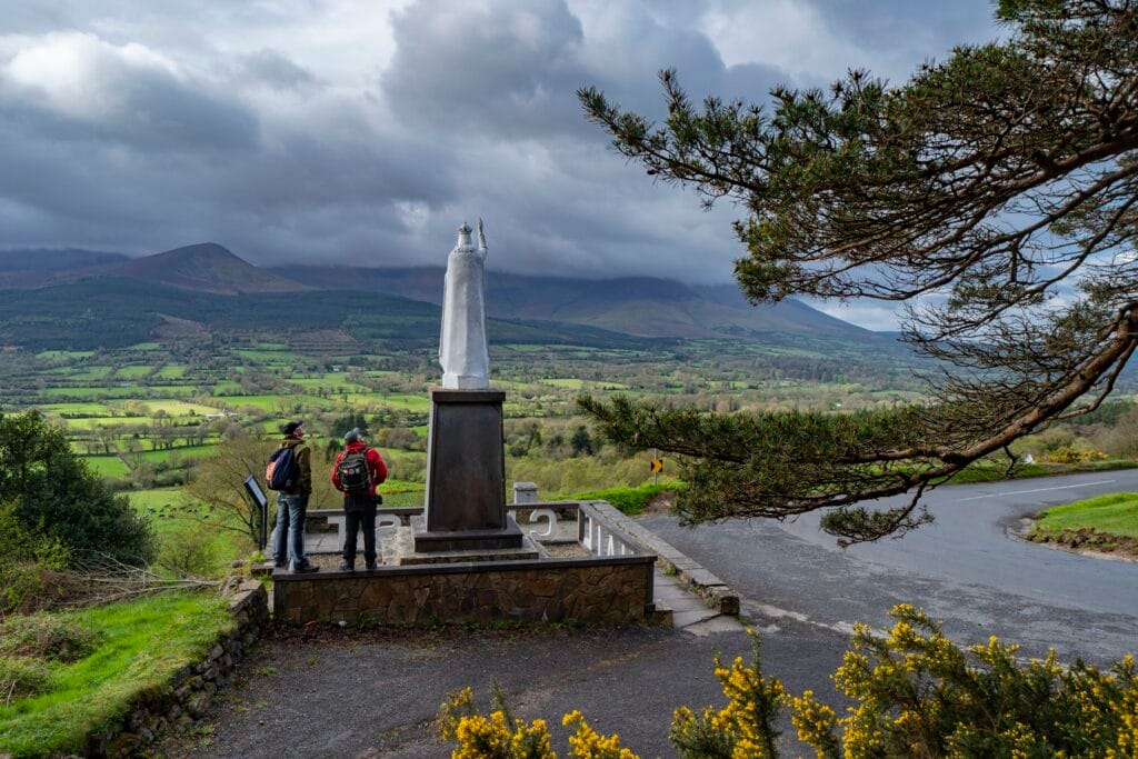 Two men looking up at the white Christ The King monument a top the side of Glen of Aherlow, with the sweeping clouds and landscape below overlooking the panoramic viewpoint in one of the world’s top destinations, Tipperary.