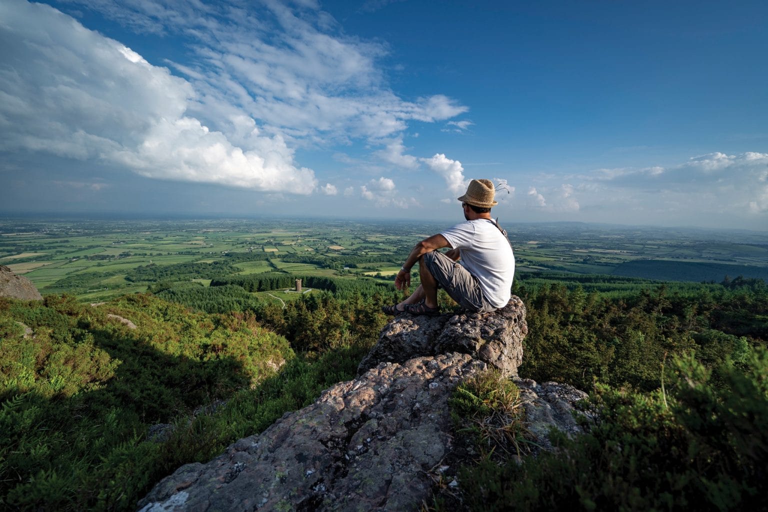 A person wearing a straw hat, white t-shirt, and shorts sits on a rocky cliff overlooking a vast green landscape with scattered trees, fields, and a distant tower under a partly cloudy sky.