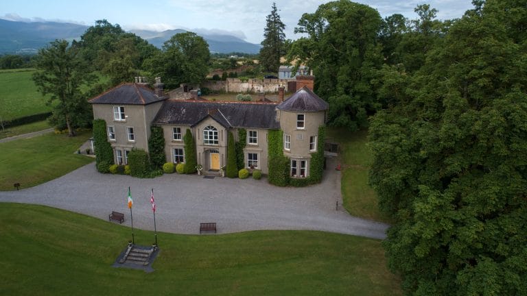 Aerial view of a large traditional mansion surrounded by green lawns, trees, and a gravel driveway, with two flagpoles displaying the Irish and English flags in front of the house, set against a backdrop of distant mountains under a partly cloudy sky.