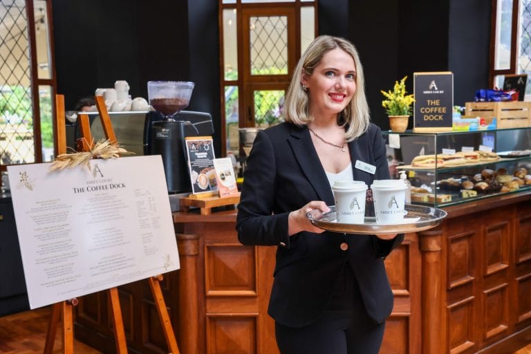 Three people sitting around a wicker outdoor table in a garden, enjoying drinks and a spread of cheese, grapes, and wine, with greenery and trees in the background under a cloudy sky.