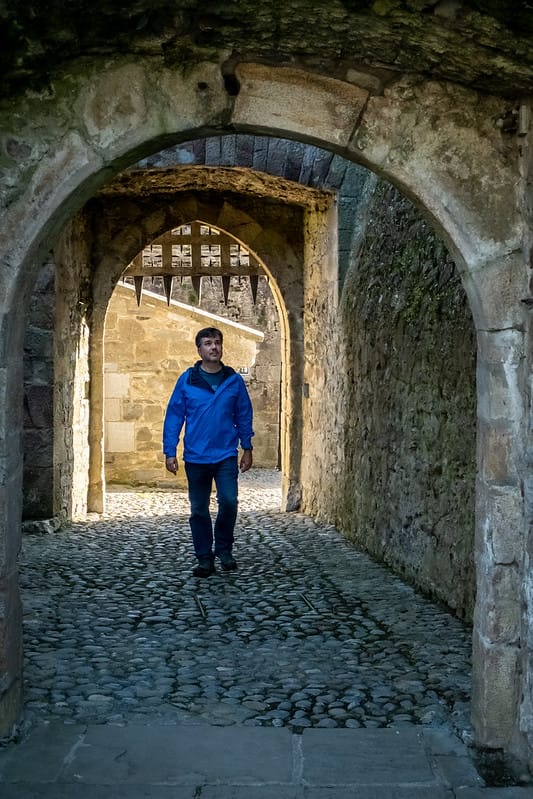 A man wearing a blue jacket walks through a stone archway in an old fortress or castle passage with cobblestone ground and a wooden portcullis visible in the background.