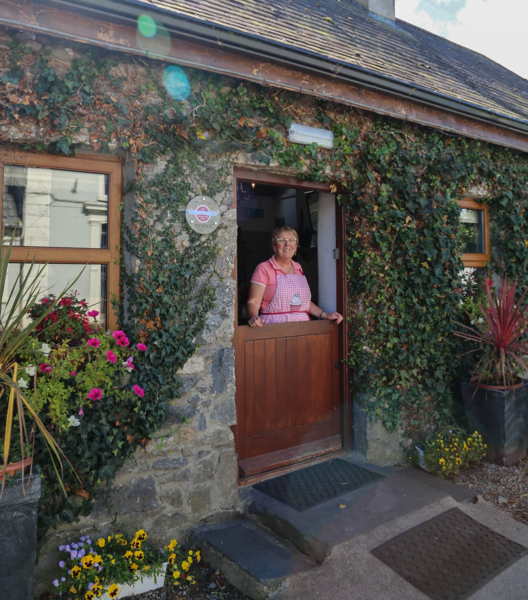 Woman in a pink checkered apron standing behind a half-door of a stone cottage covered in green ivy and surrounded by colorful flowers.