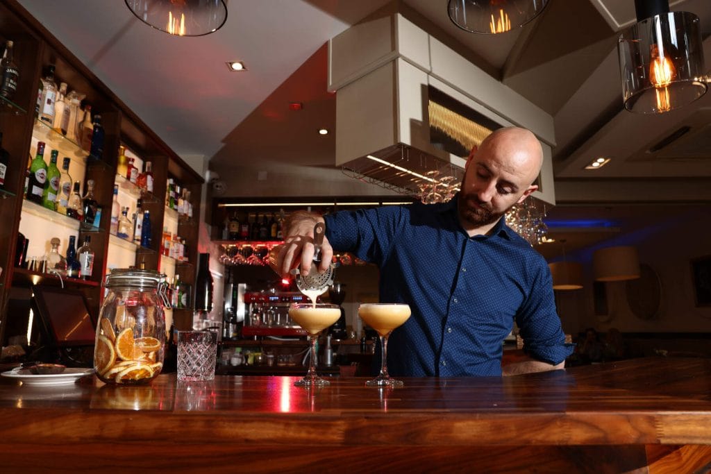 Bartender in a blue shirt pouring a cocktail through a strainer into a glass at a wooden bar counter in a dimly lit bar with shelves of liquor bottles and hanging glasses in the background.