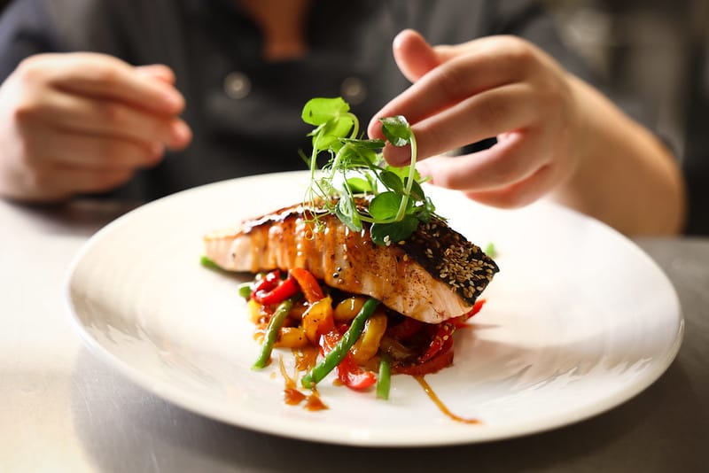 A close-up of a cooked salmon fillet garnished with sesame seeds and fresh greens, placed on a bed of sautéed mixed vegetables, being carefully arranged by a person’s hands on a white plate.