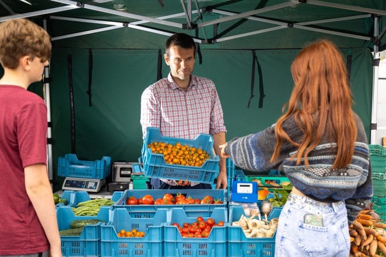 A man in a plaid shirt stands behind a market stall with blue crates filled with vegetables, handing a crate of cherry tomatoes to a woman with long red hair pointing at the produce, while a boy in a maroon shirt stands nearby.