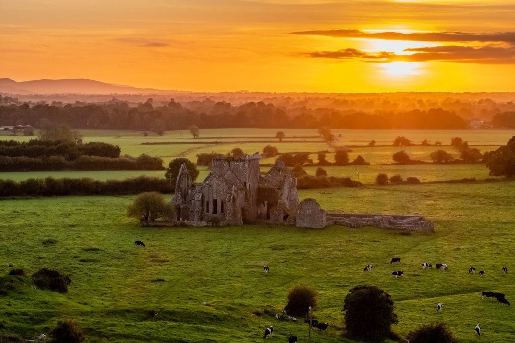 Sunset over a green countryside with a historic stone ruin in the center and cows grazing in the field.