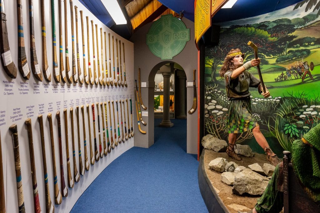 Museum exhibit featuring a display of numerous vintage hurling sticks mounted on a curved wall with labels, and a lifelike statue of a medieval warrior holding a hurley in a battle scene with a painted backdrop of a historic landscape and warriors.