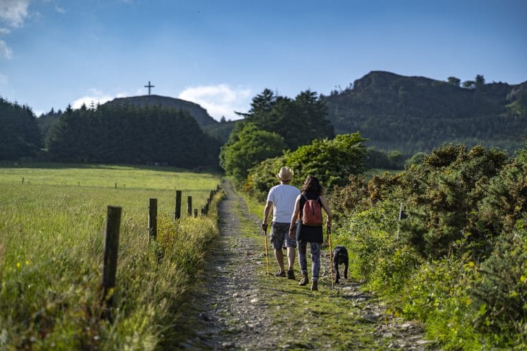 Two hikers with walking sticks and a black dog walk on a forest path toward a hilltop with a large cross under a blue sky.