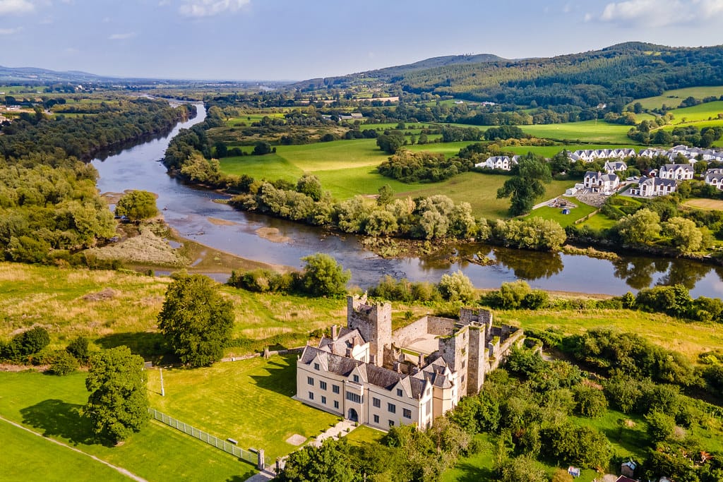 Aerial view of a historic castle near a winding river surrounded by lush green fields and trees, with a small residential area and hills in the distance under a partly cloudy sky.